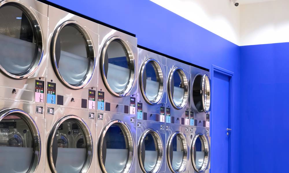Front-load washing machines are stacked on top of each other in front of a dark blue wall at a laundromat.