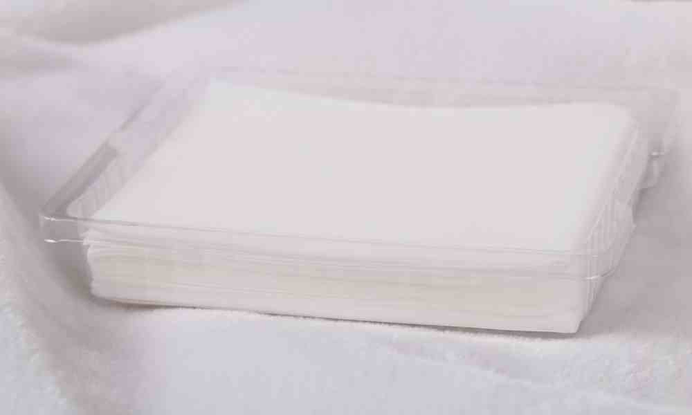 A short stack of white dryer sheets sits inside a clear plastic container on top of a soft, white blanket.